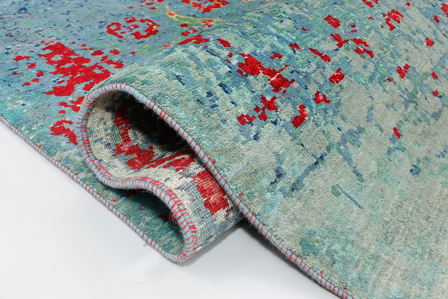 A close-up of a transitional rug showcasing precise knotting, where a light-catching hybrid silk blend illuminates a textured, watery mix of blue and teal yarns surrounding a faded crimson geometric motif.