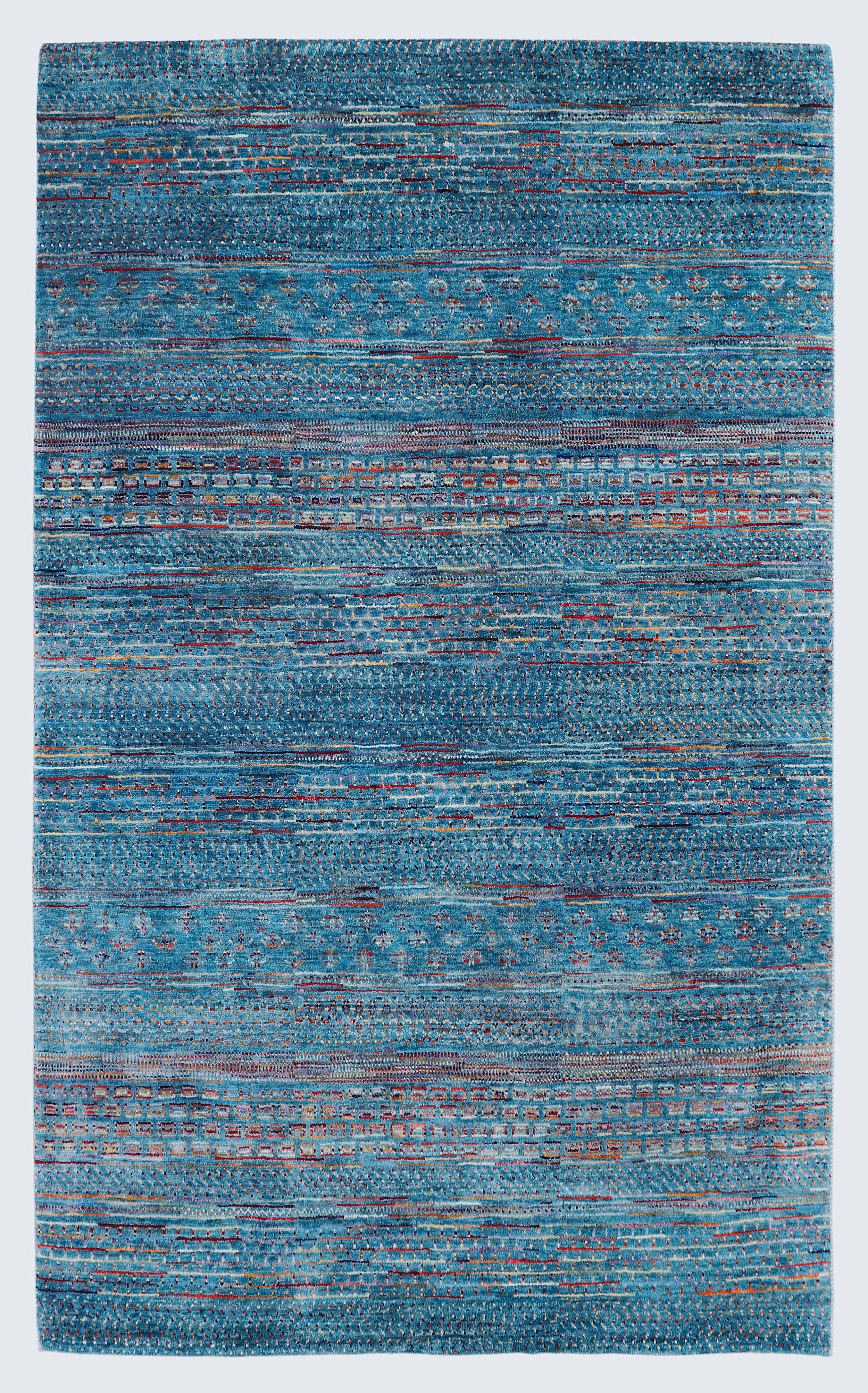 A full view of a vibrant hand-knotted Gabbeh rug from the Aatish collection, featuring horizontal bands of gold, rust, white, and green tribal motifs over a striated azure-blue foundation.