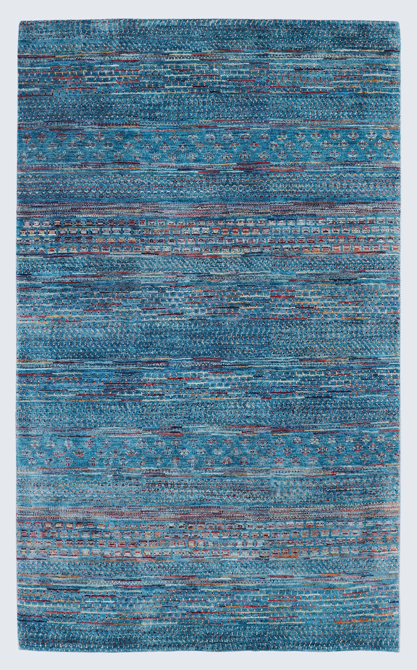 A full view of a vibrant hand-knotted Gabbeh rug from the Aatish collection, featuring horizontal bands of gold, rust, white, and green tribal motifs over a striated azure-blue foundation.