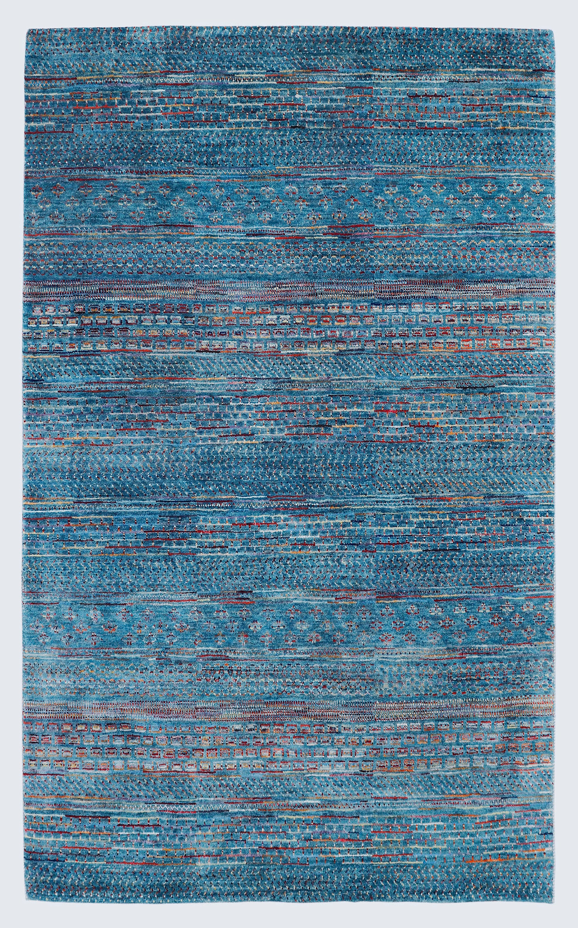 A full view of a vibrant hand-knotted Gabbeh rug from the Aatish collection, featuring horizontal bands of gold, rust, white, and green tribal motifs over a striated azure-blue foundation.