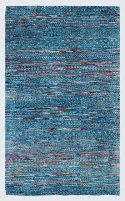 A full view of a vibrant hand-knotted Gabbeh rug from the Aatish collection, featuring horizontal bands of gold, rust, white, and green tribal motifs over a striated azure-blue foundation.