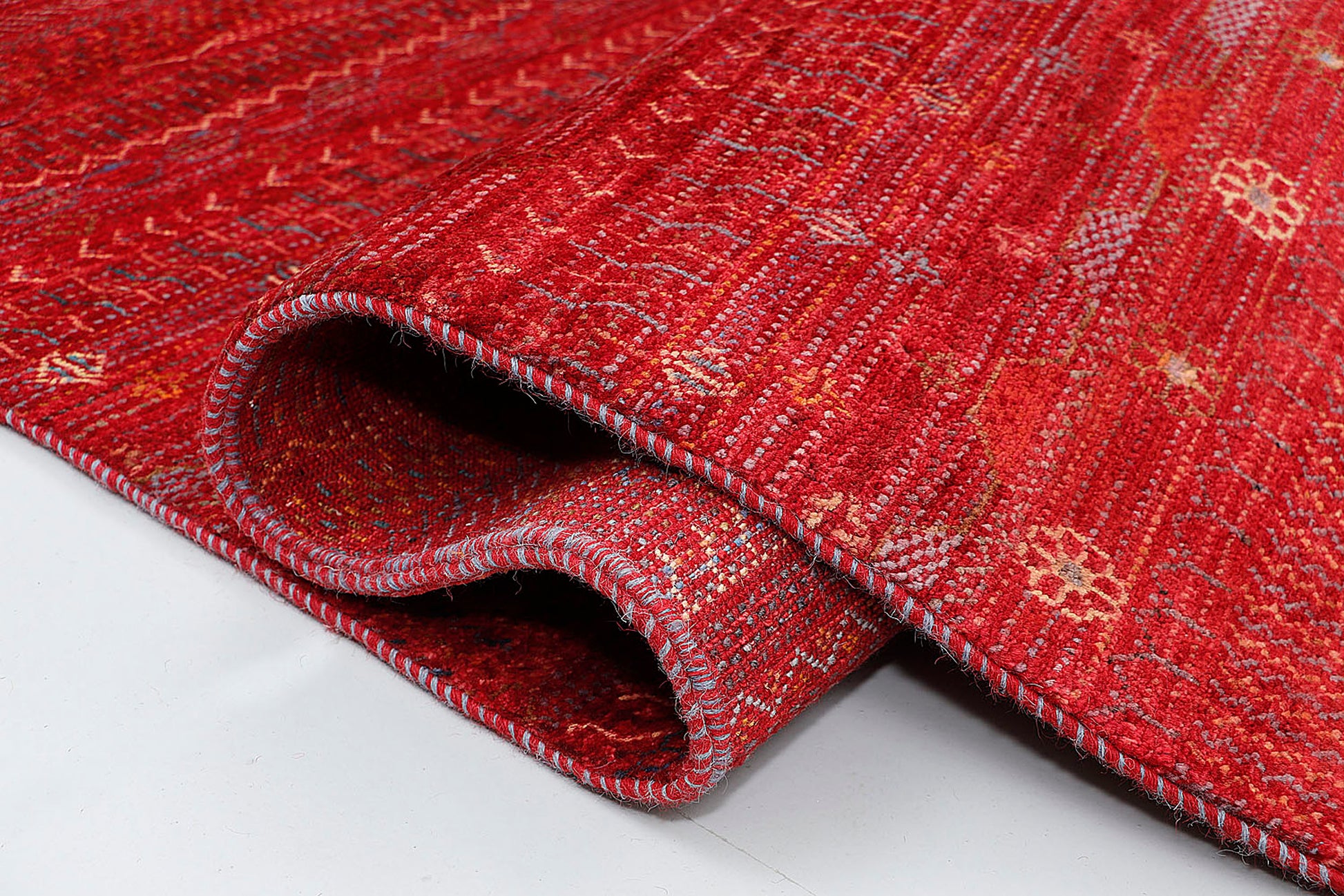 A close-up of a thick-pile wool rug highlighting the dense, hand-knotted texture and the rhythmic, script-like rows of colorful nomadic symbols against a rich red background.