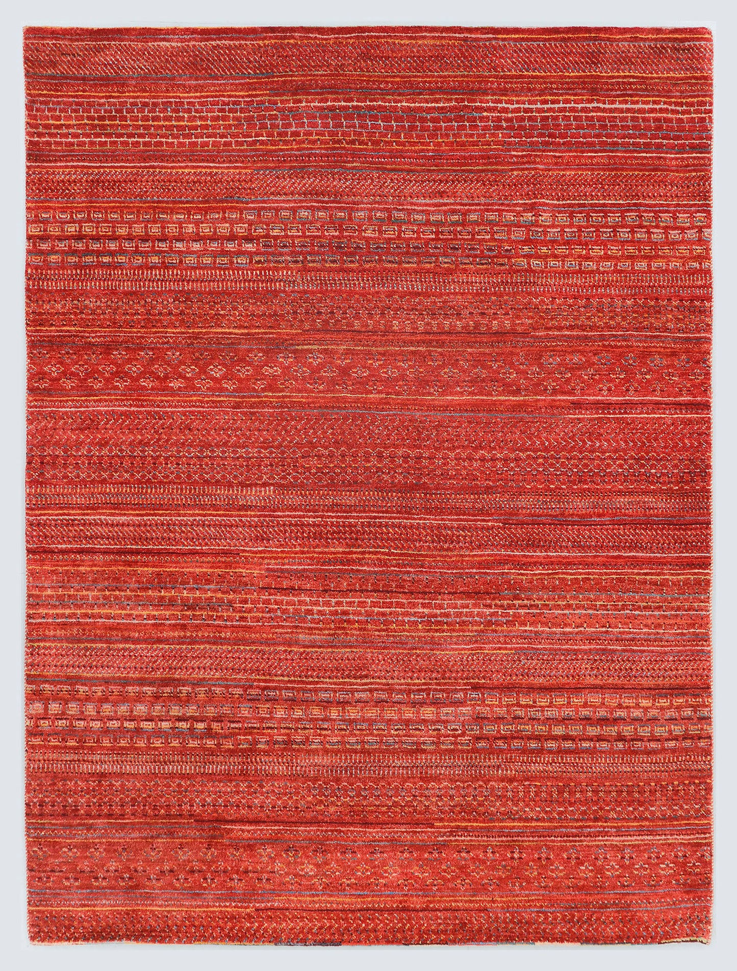 A full view of an earthy, hand-knotted Gabbeh rug from the Aatish collection, featuring horizontal bands of gold and slate blue tribal motifs over a textured copper-rust foundation.