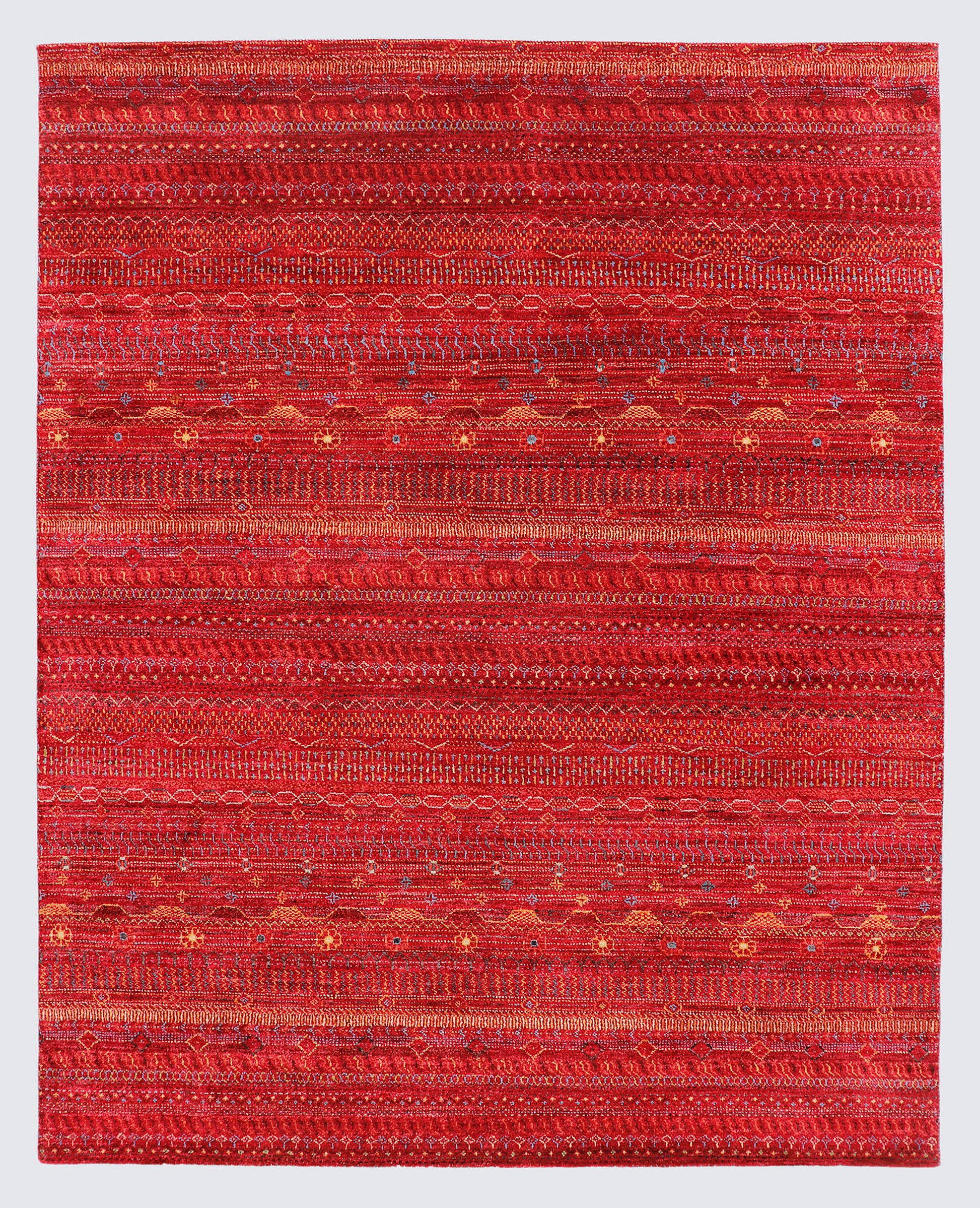 A full view of an earthy, hand-knotted Gabbeh rug from the Aatish collection, featuring horizontal bands of gold, orange, and teal tribal motifs over a textured rust-red foundation.