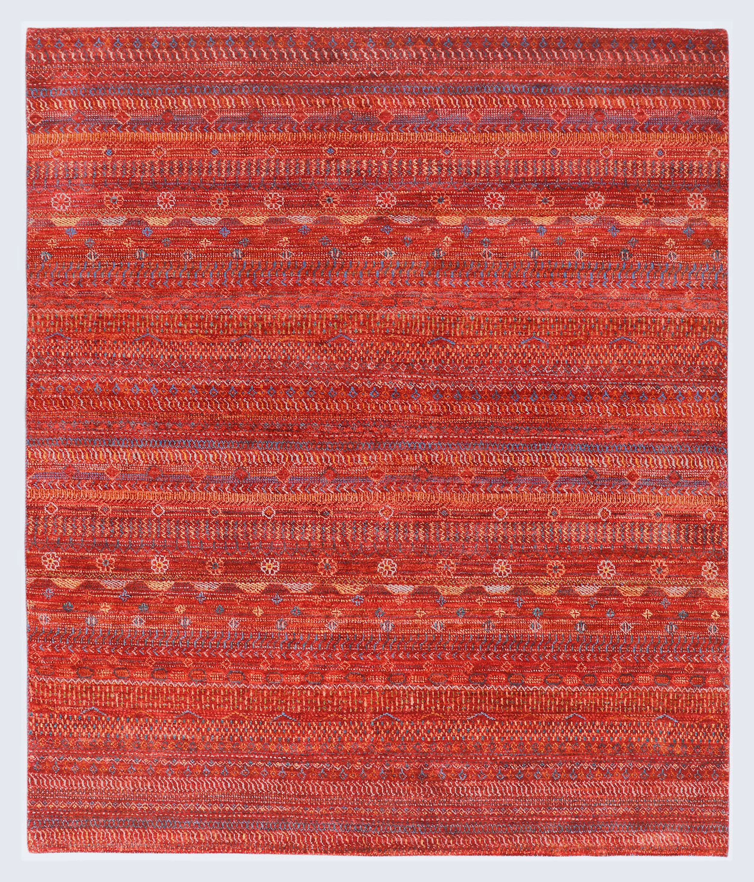 A full view of a warm, hand-knotted Gabbeh rug from the Aatish collection, featuring horizontal bands of gold, blue, white, and green tribal motifs over a heavily textured rust-orange foundation.