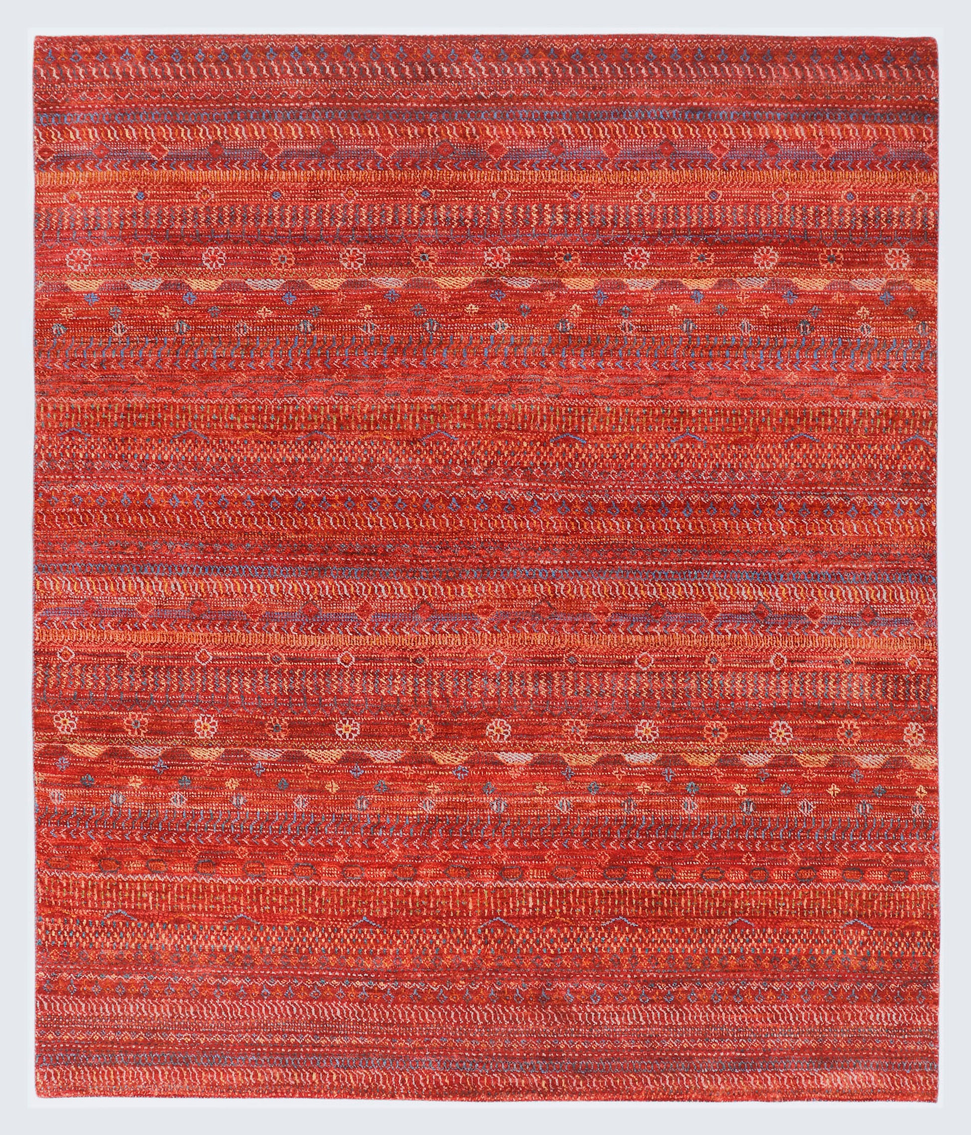 A full view of a warm, hand-knotted Gabbeh rug from the Aatish collection, featuring horizontal bands of gold, blue, white, and green tribal motifs over a heavily textured rust-orange foundation.