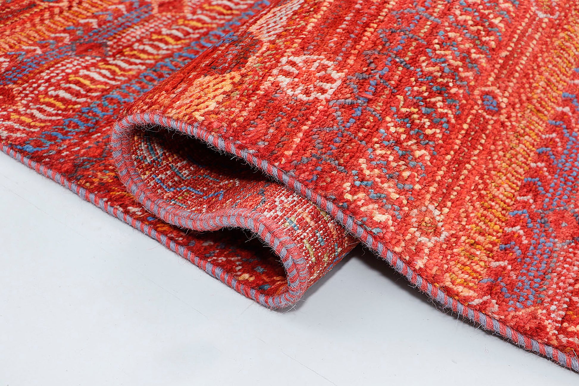 A close-up of a thick-pile wool rug highlighting the plush, organic texture and the vibrant, multi-colored geometric motifs woven into the earthy russet-red base.
