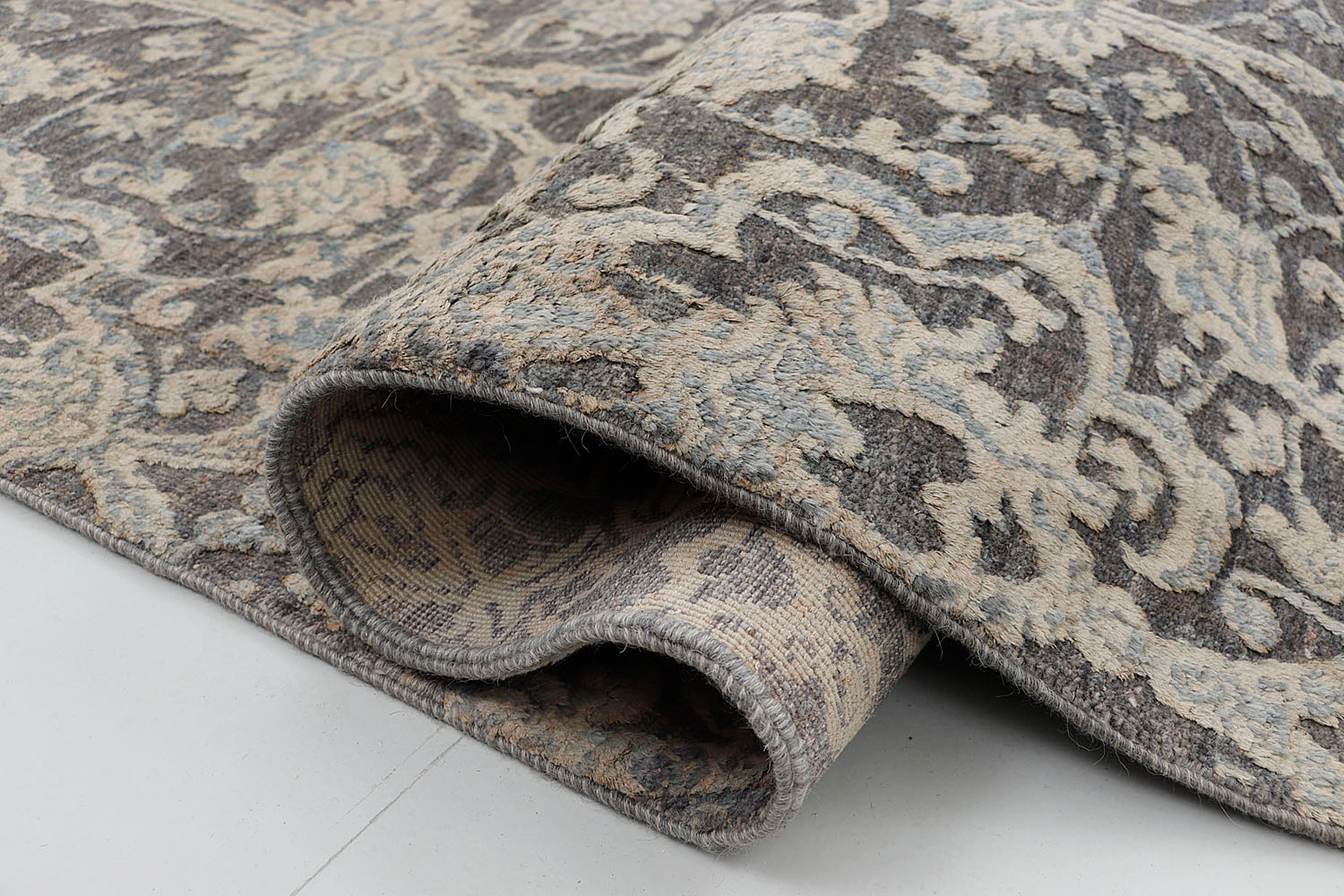 A close-up of a sculpted hand-knotted wool rug highlighting the high-low pile technique, showing the raised classical motifs creating physical depth against a hand-carved dark grey foundation.